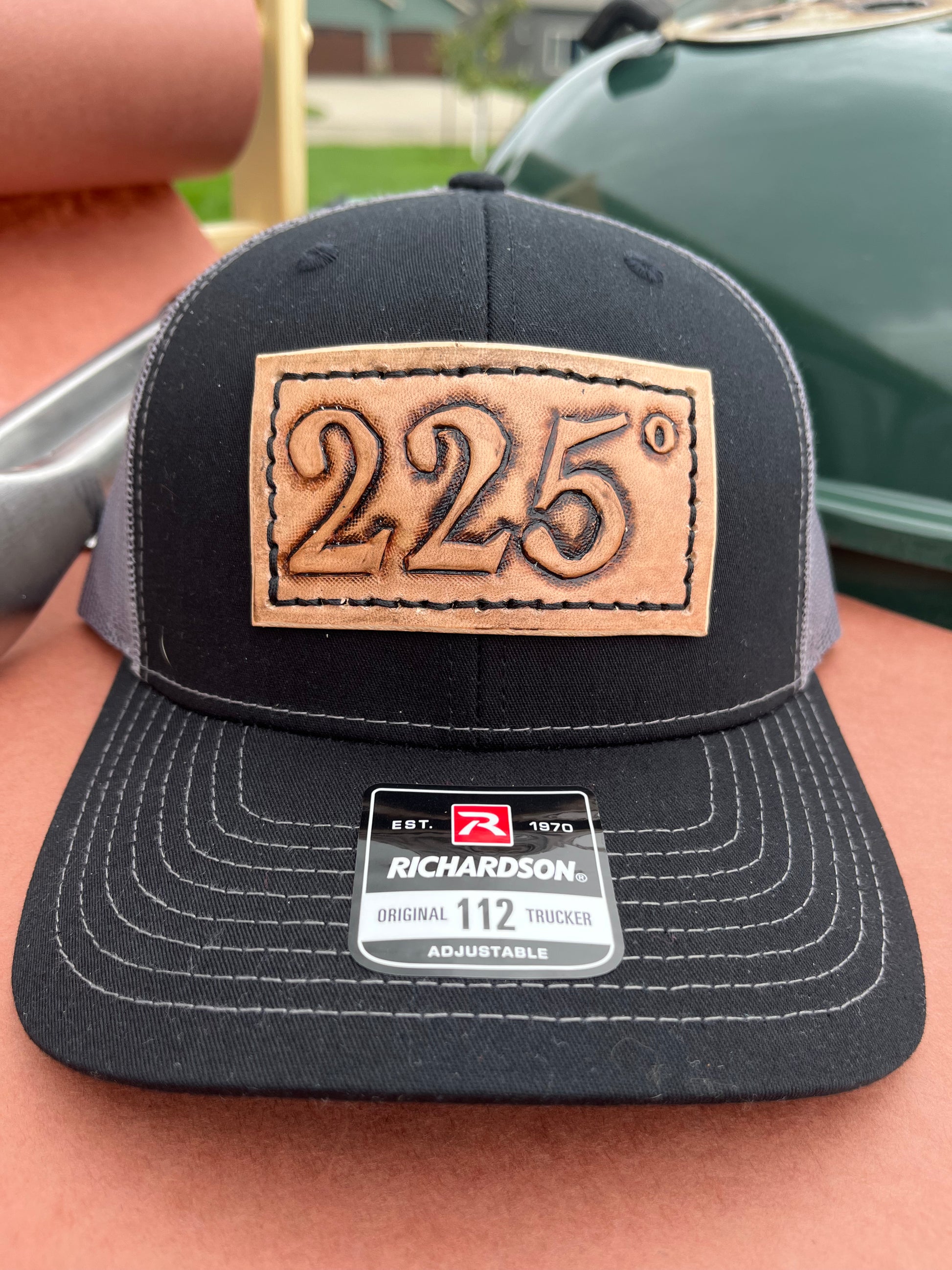 Black and gray richardson 112 trucker hat with a hand stitched leather patch on the front with 225 degrees tooled into the patch